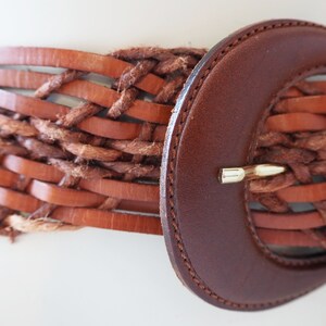 May include: A brown leather belt with a woven design and a large, brown leather buckle.
