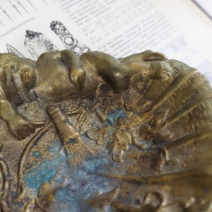 May include: A vintage brass ashtray with a detailed design of a reclining figure and other decorative elements. The ashtray has a worn, aged patina with hints of green and blue.