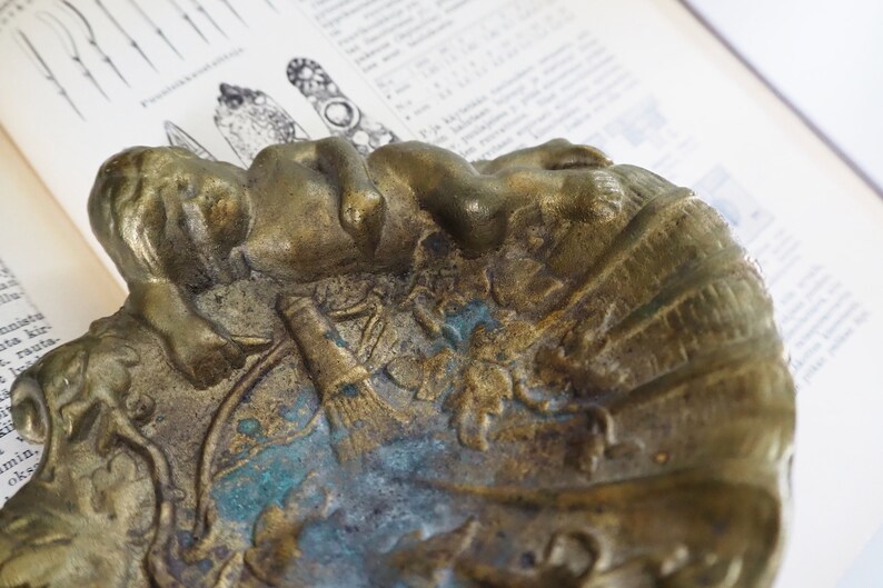 May include: A vintage brass ashtray with a detailed design of a reclining figure and other decorative elements. The ashtray has a worn, aged patina with hints of green and blue.