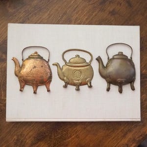 May include: Three decorative metal teapots in gold and copper tones, mounted on a white background. The teapots have a traditional design with a rounded body and a spout.