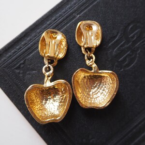 May include: A pair of gold-tone clip-on earrings shaped like apples. The earrings have a textured surface and are dangling from a small gold-tone loop.