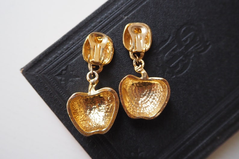 May include: A pair of gold-tone clip-on earrings shaped like apples. The earrings have a textured surface and are dangling from a small gold-tone loop.