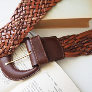 May include: A brown leather belt with a woven design and a large, brown leather buckle.