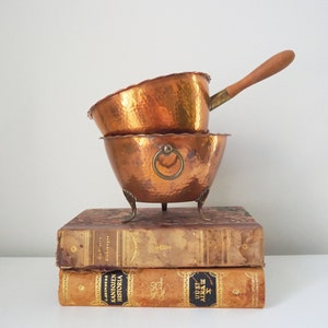 Può includere: Due pentole di rame con superfici martellate e un manico in legno. Le pentole sono impilate l'una sull'altra e sono appoggiate su due libri. I libri sono marroni e hanno scritte dorate sulle copertine.
