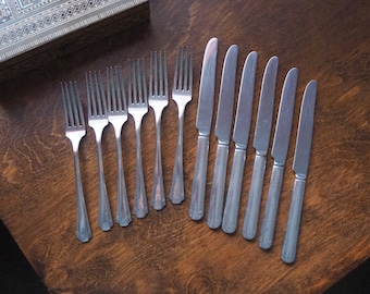 May include: A collection of vintage cutlery, comprising six forks and six knives, displayed on a wooden surface. The silverware is silver-toned with ornate handle designs. The wooden surface has a warm, brown colour.