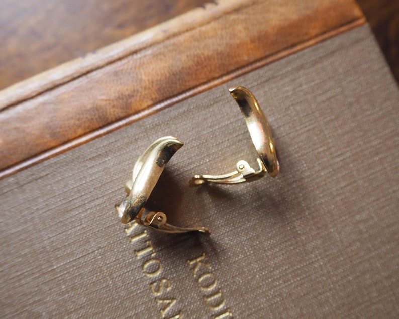May include: A pair of gold-toned clip-on earrings. The earrings are shaped like a crescent moon and have a simple, elegant design.
