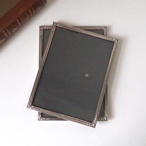 May include: Two rectangular silver picture frames with decorative corner detailing are stacked on a white surface. The frames have dark grey backing. A vintage brown book is in the upper left corner.