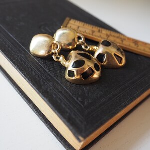 May include: A pair of gold tone earrings with black geometric shapes. The earrings are dangling and have a round top.