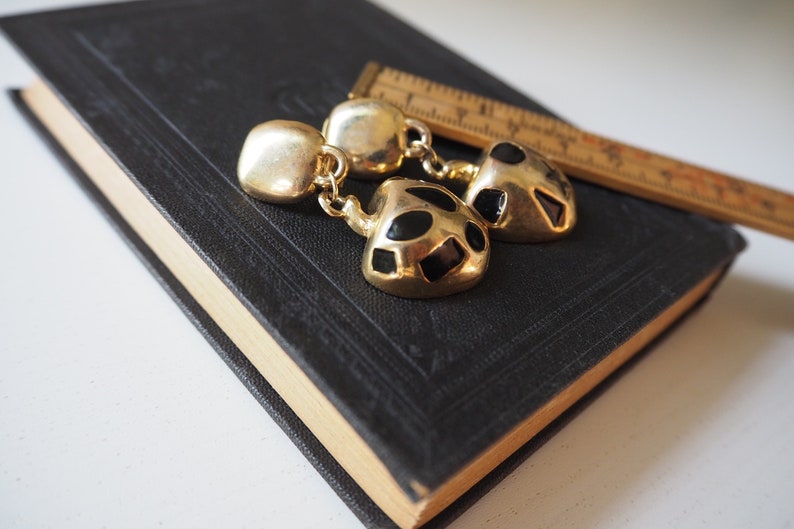 May include: A pair of gold tone earrings with black geometric shapes. The earrings are dangling and have a round top.