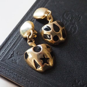 May include: A pair of gold-tone earrings with black enamel accents. The earrings are shaped like apples with a star and polka dots on one and a polka dot pattern on the other.