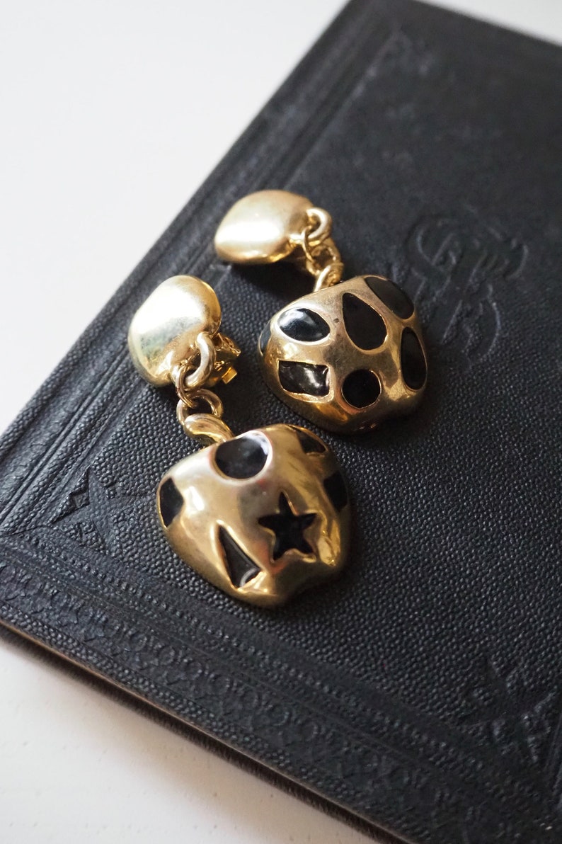 May include: A pair of gold-tone earrings with black enamel accents. The earrings are shaped like apples with a star and polka dots on one and a polka dot pattern on the other.