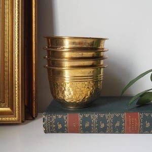 May include: A stack of four antique brass bowls with a hammered texture on the bottom bowl. The bowls are stacked on a dark blue book with gold lettering. A framed painting is in the background.