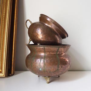 Peut inclure: Deux pots de fleurs en cuivre avec une texture martelée. Le plus petit pot est empilé sur le plus grand. Les deux pots ont des poignées et des pieds en laiton.