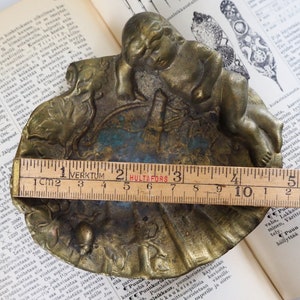 May include: A brass trinket dish with a sleeping cherub figure on top. The dish is decorated with a floral design and has a blue and green patina. The dish measures approximately 4 inches in diameter.
