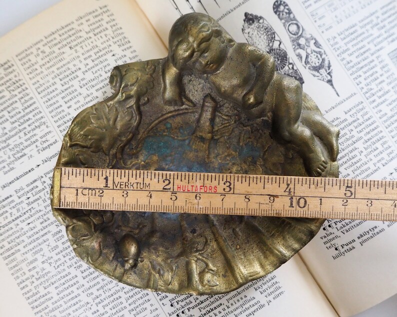 May include: A brass trinket dish with a sleeping cherub figure on top. The dish is decorated with a floral design and has a blue and green patina. The dish measures approximately 4 inches in diameter.