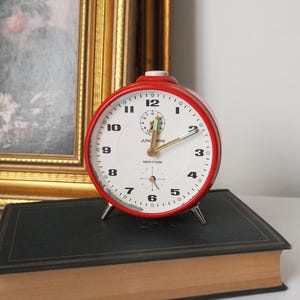 May include: Red alarm clock with white face and black numbers. The clock has a gold hand and says "JUNGHANS REPETITION" on the face.