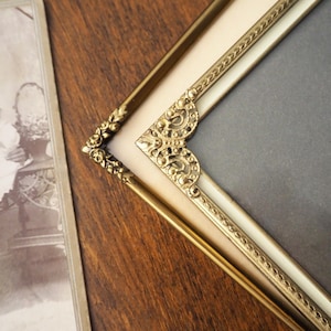 Könnte beinhalten: Nahaufnahme eines Vintage-Bilderrahmens in Goldoptik mit verzierten Eckdetails. Der Rahmen hat einen cremefarbenen Innenrand und einen dunkelgrauen Hintergrund. Auf der linken Seite ist eine Sepia-Fotografie teilweise sichtbar, die auf einer Holzoberfläche liegt.