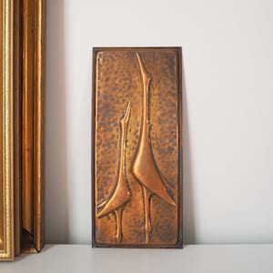 Pode incluir: Uma peça de arte de parede retangular de cobre com duas aves estilizadas de pescoço comprido. As aves são levantadas contra um fundo texturizado. A obra de arte é emoldurada em uma borda marrom escura.