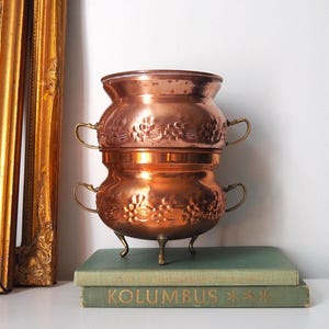 Può includere: Due pentole di rame impilate, con motivi floreali e piccole maniglie. La pentola inferiore ha tre piccole gambe. Le pentole sono appoggiate su libri verdi. Uno specchio con cornice dorata sullo sfondo. Diametro circa 20 cm.