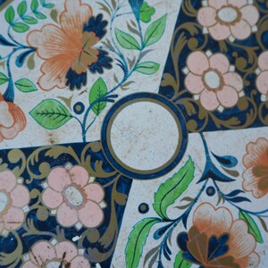 Puede incluir: Una bandeja de hojalata vintage con fondo blanco y dise&ntilde;o floral azul y dorado. La bandeja tiene un centro circular con fondo blanco y borde dorado.