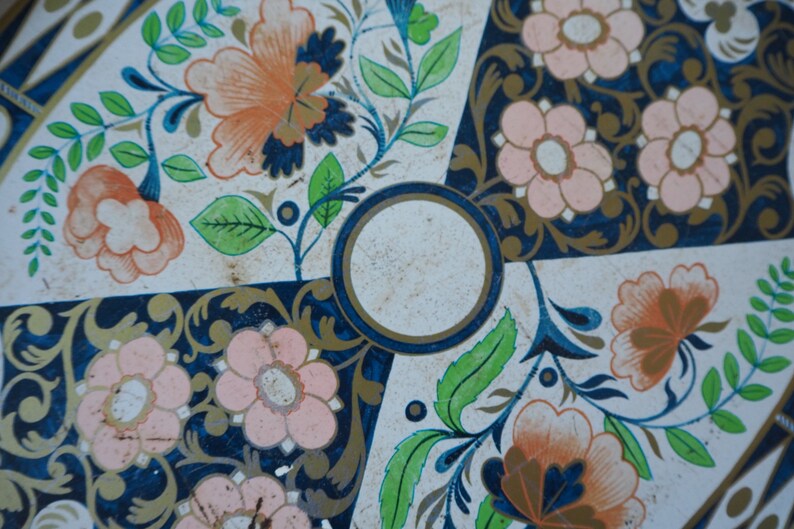 Puede incluir: Una bandeja de hojalata vintage con fondo blanco y dise&ntilde;o floral azul y dorado. La bandeja tiene un centro circular con fondo blanco y borde dorado.