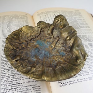 May include: A vintage brass trinket dish with an ornate design featuring a cherub figure. The dish has a scalloped edge and a textured surface.