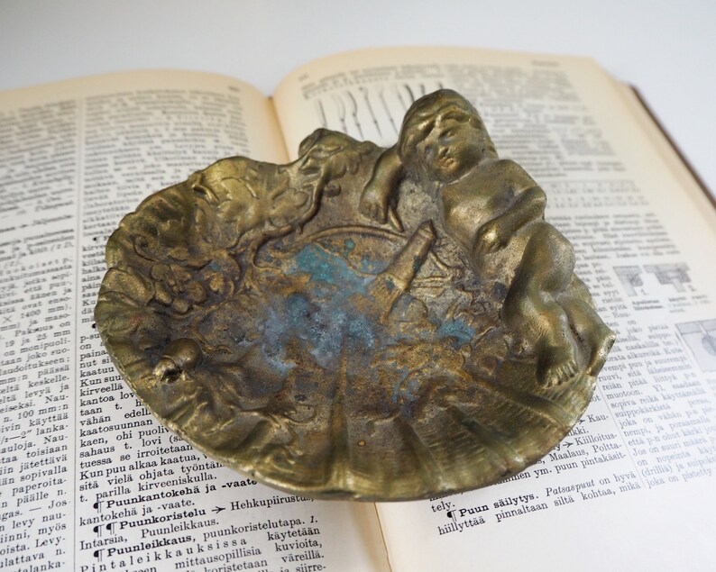 May include: A vintage brass trinket dish with an ornate design featuring a cherub figure. The dish has a scalloped edge and a textured surface.