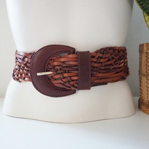 May include: A brown leather braided belt with a large, round, brown buckle. The belt is woven with a variety of brown leather strips.