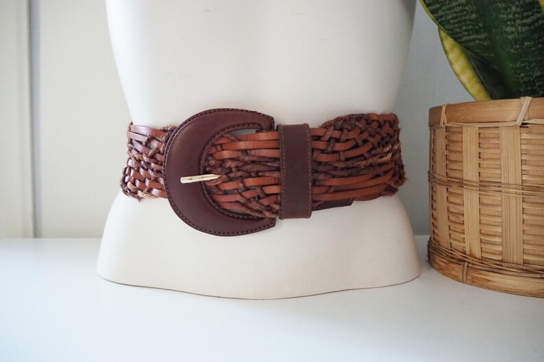 May include: A brown leather braided belt with a large, round, brown buckle. The belt is woven with a variety of brown leather strips.