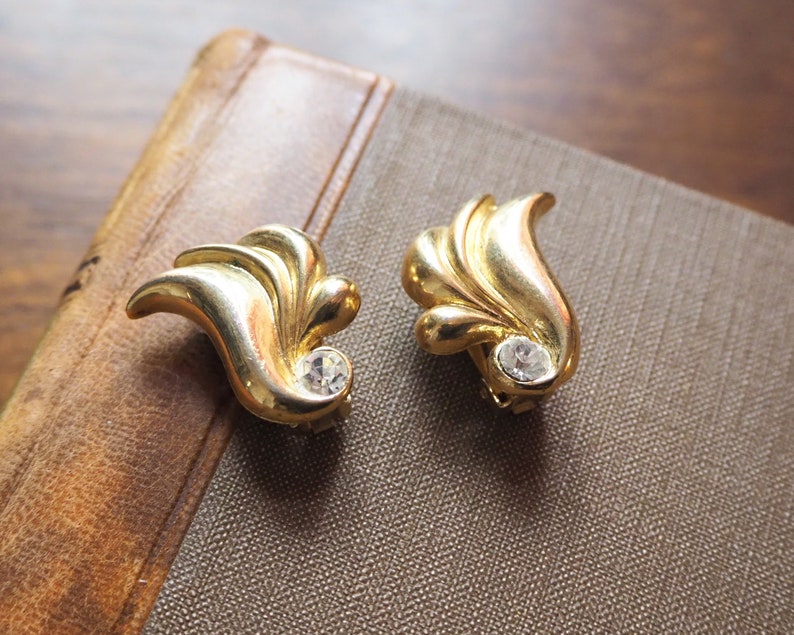 May include: A pair of gold-tone earrings with a leaf design and a clear rhinestone in the center of each earring.