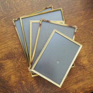 Könnte beinhalten: Eine Sammlung antiker goldfarbener Bilderrahmen mit verzierten Eckdetails. Die rechteckigen Rahmen sind auf einer Holzoberfläche angeordnet. Die Rahmen haben eine dunkelgraue Rückseite.