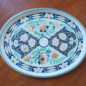 Puede incluir: Una bandeja de servir ovalada de metal con fondo blanco y un dise&ntilde;o floral en tonos azules, rosas y verdes. La bandeja tiene un borde dorado.