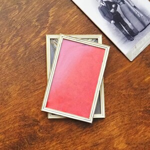 Puede incluir: Dos marcos de fotos rectangulares con borde dorado, uno con fondo rojo y otro con fondo gris, apilados sobre una superficie de madera. Una fotografía vintage en blanco y negro descansa cerca.