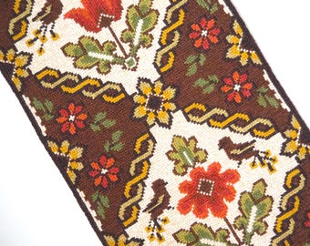 Op de afbeelding: Close-up van een geweven textiel met een herhalend bloemenpatroon. Het ontwerp toont rode, oranje en gele bloemen, groene bladeren en bruine vogels op een crème- en bruine achtergrond. De rand heeft een geel en bruin kettingachtig ontwerp.