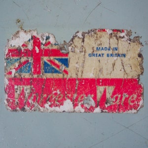 Puede incluir: Una etiqueta vintage roja, blanca y azul con el texto "Made in Great Britain" y una imagen descolorida de la bandera de la Union Jack. La etiqueta est&aacute; parcialmente rasgada y tiene un fondo rojo con la palabra "Ware" en blanco.
