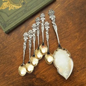 Puede incluir: Un conjunto de cucharas de servir y una espátula de plata antigua, con mangos en forma de rosa y cuencos dorados. Las cucharas están dispuestas sobre una superficie de madera junto a un libro verde.
