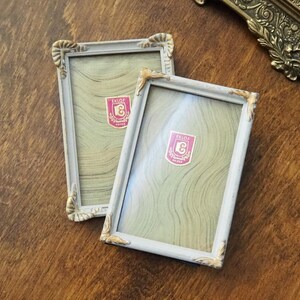Könnte beinhalten: Zwei rechteckige Vintage-Bilderrahmen mit weißem Antik-Finish und goldfarbenen Eckakzenten. Jeder Rahmen hat ein rosa-weißes Etikett mit dem Text "EKLOF". Die Rahmen liegen auf einer Holzoberfläche.