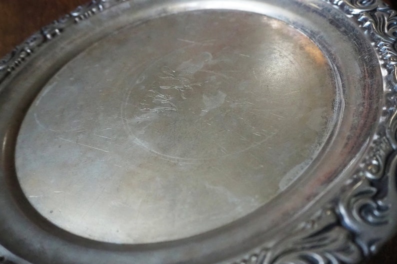 May include: A silver serving tray with a decorative, ornate rim. The tray has a smooth, reflective surface with a faint design in the center.