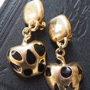 May include: A pair of gold-tone clip-on earrings featuring black enamel apple charms with black polka dots. One apple charm has a star on it.