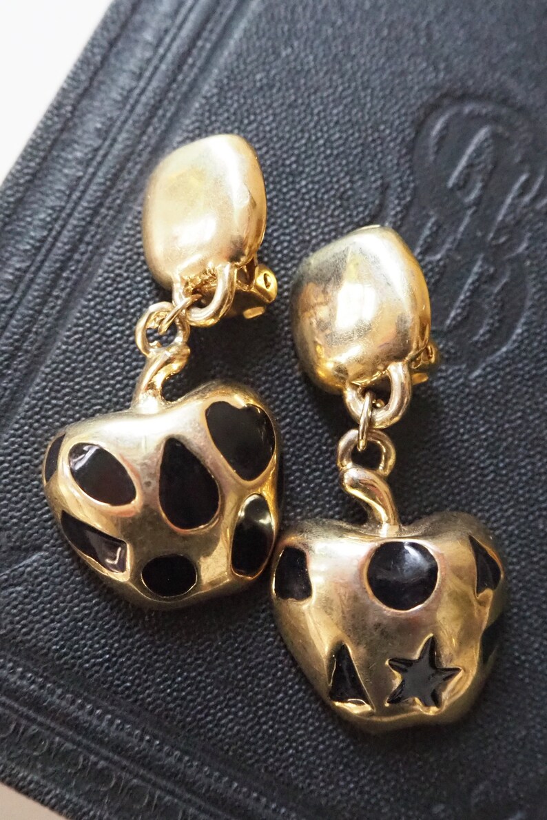 May include: A pair of gold-tone clip-on earrings featuring black enamel apple charms with black polka dots. One apple charm has a star on it.