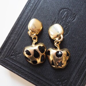 May include: A pair of gold-tone dangle earrings with black enamel accents. The earrings feature a heart-shaped design with black dots and a star.