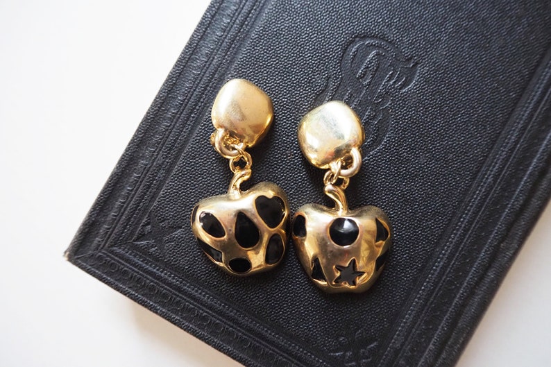May include: A pair of gold-tone dangle earrings with black enamel accents. The earrings feature a heart-shaped design with black dots and a star.