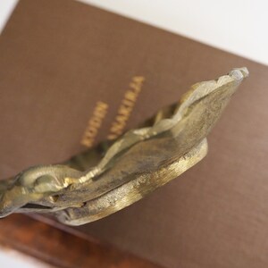 May include: A gold-toned metal bookend with an ornate design. The bookend is shaped like a stylized leaf with a raised edge.