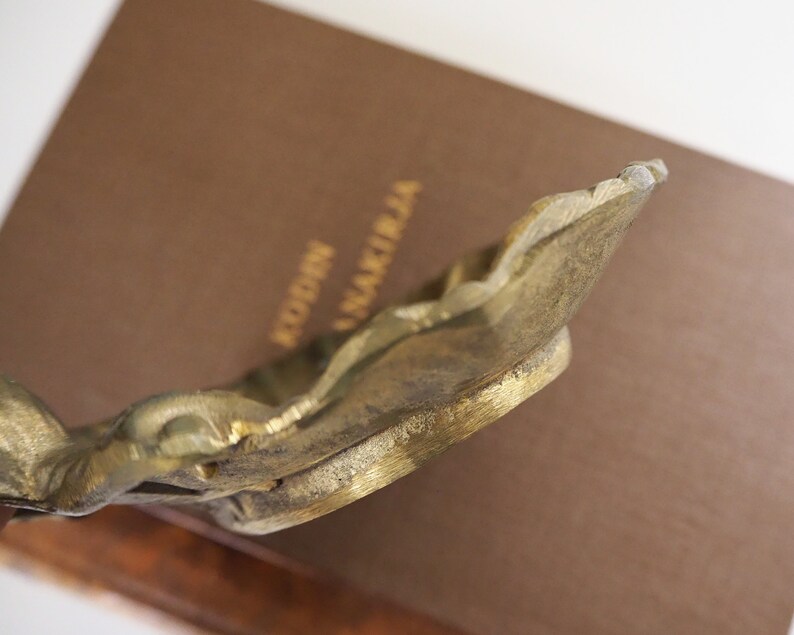 May include: A gold-toned metal bookend with an ornate design. The bookend is shaped like a stylized leaf with a raised edge.