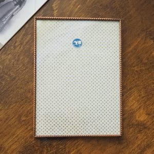 Puede incluir: Un marco de fotos rectangular de color cobre con un borde retorcido en forma de cuerda. El marco contiene un papel estampado con pequeños puntos grises. Una fotografía en blanco y negro es parcialmente visible en la esquina superior izquierda.
