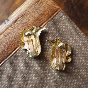 May include: A pair of gold-tone clip-on earrings with a floral design. The earrings have a leaf-like shape with a small, round, gold-tone accent in the center.
