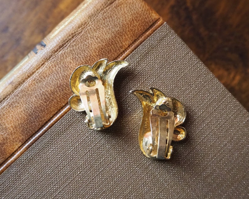 May include: A pair of gold-tone clip-on earrings with a floral design. The earrings have a leaf-like shape with a small, round, gold-tone accent in the center.