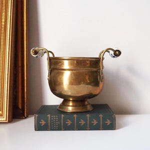 Pode incluir: Uma tigela de latão vintage com duas alças curvas, cada uma adornada com um botão decorativo transparente. A tigela está em cima de um livro verde escuro com letras douradas. Uma pintura emoldurada é visível no fundo.