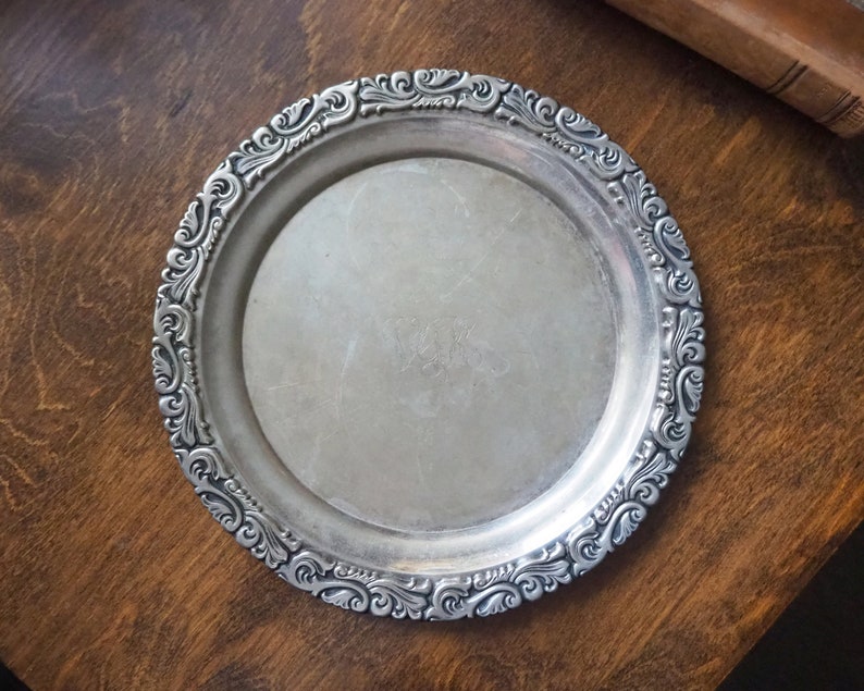 May include: A silver serving tray with a decorative, ornate border. The tray is round and has a smooth, reflective surface.