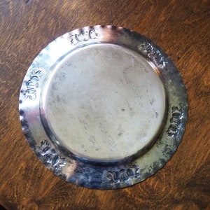 May include: A silver metal serving tray with a decorative embossed border. The tray is round and has a smooth, shiny surface.